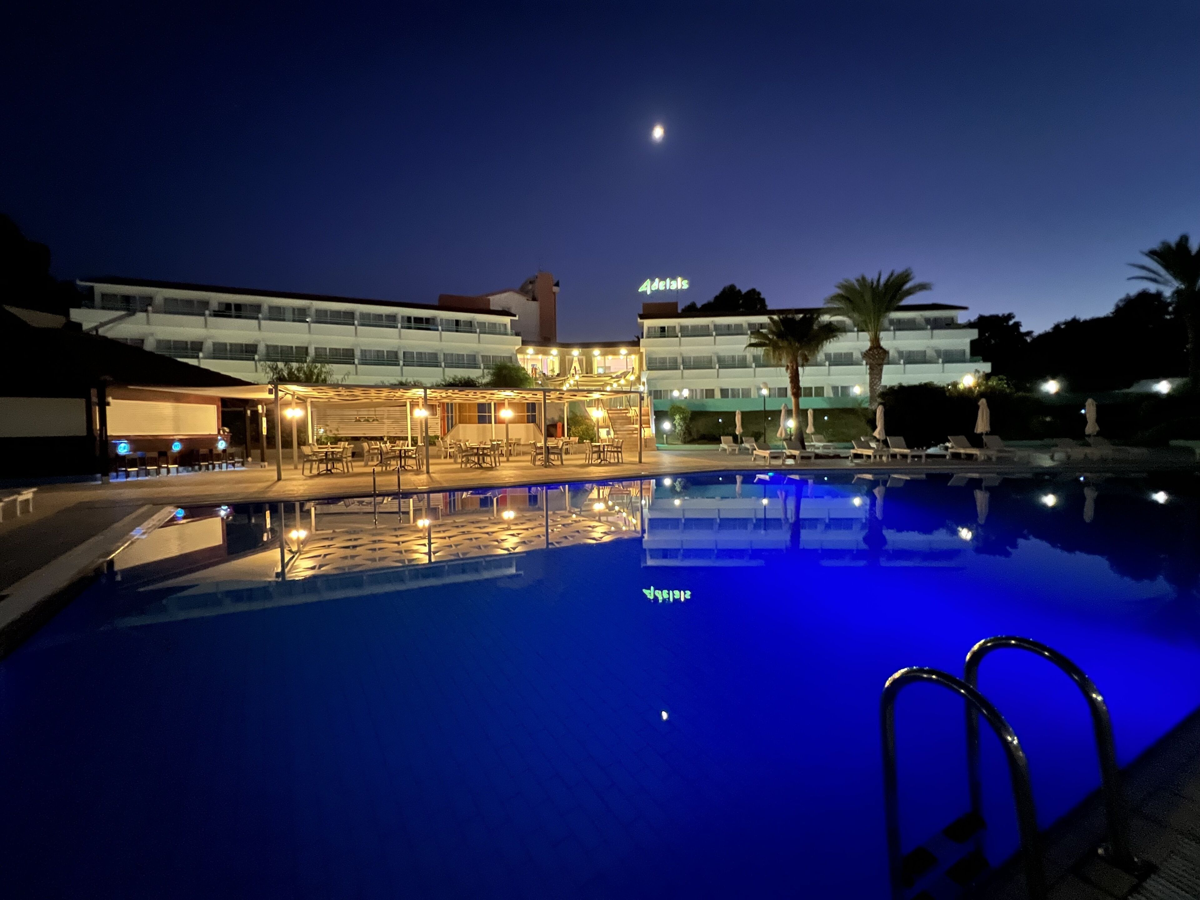 A swimming pool with lounge chairs and umbrellas, a building with a sign 'Details', and a moon in the sky.