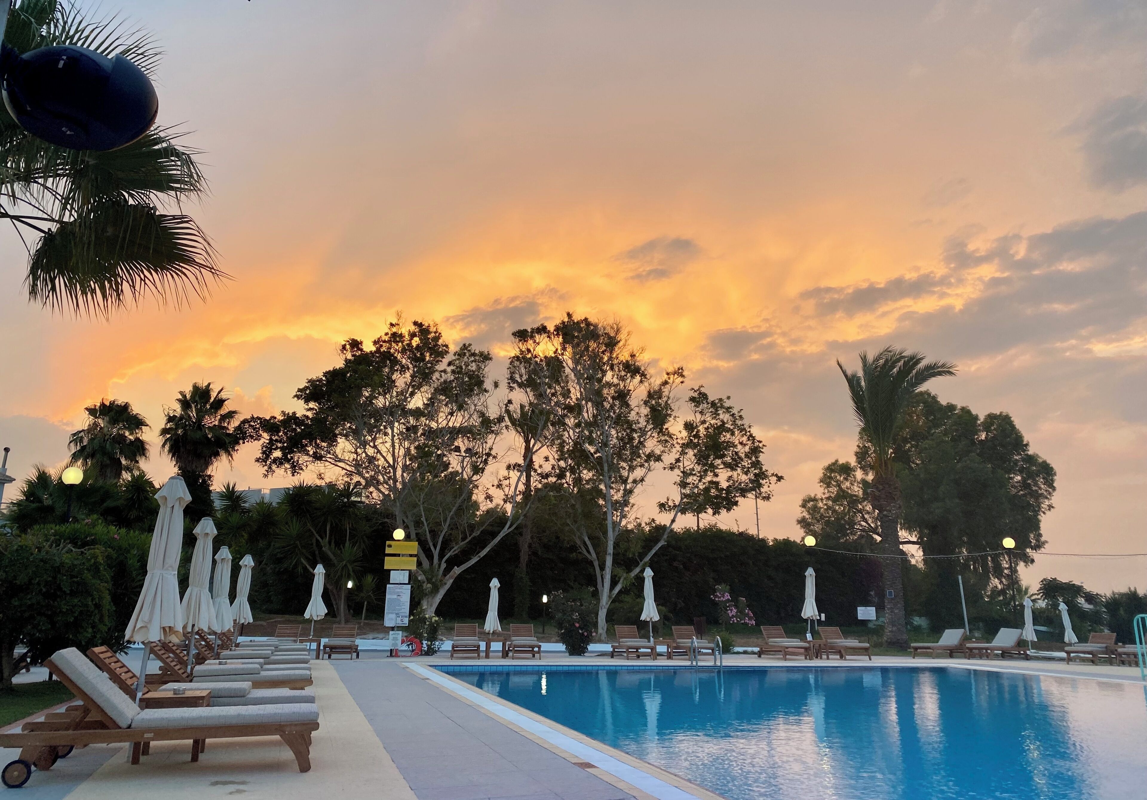 A poolside area with lounge chairs and umbrellas, surrounded by trees and a sunset sky.
