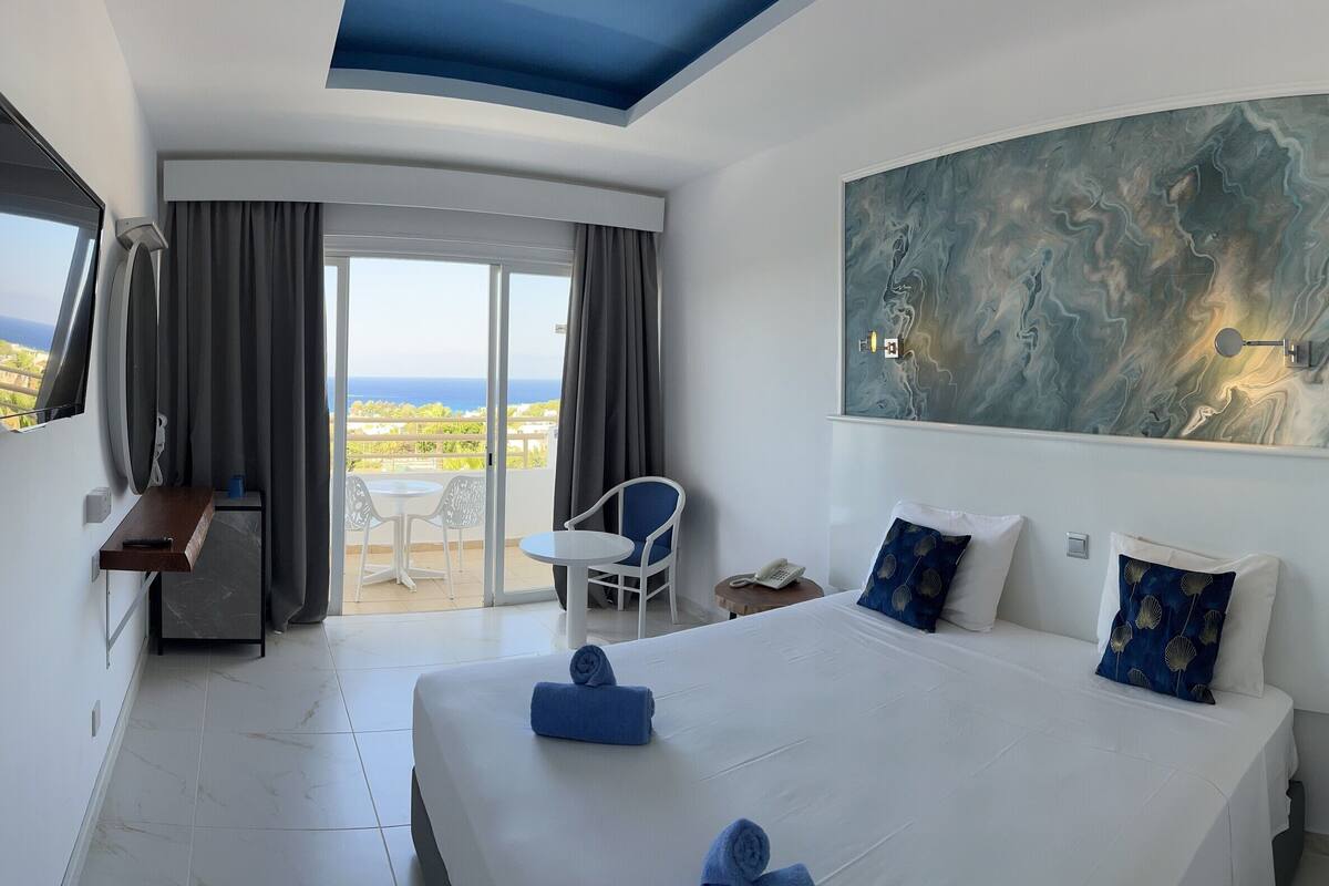 A hotel room with a large bed, a flat-screen TV, a painting on the wall, and a balcony with a view.