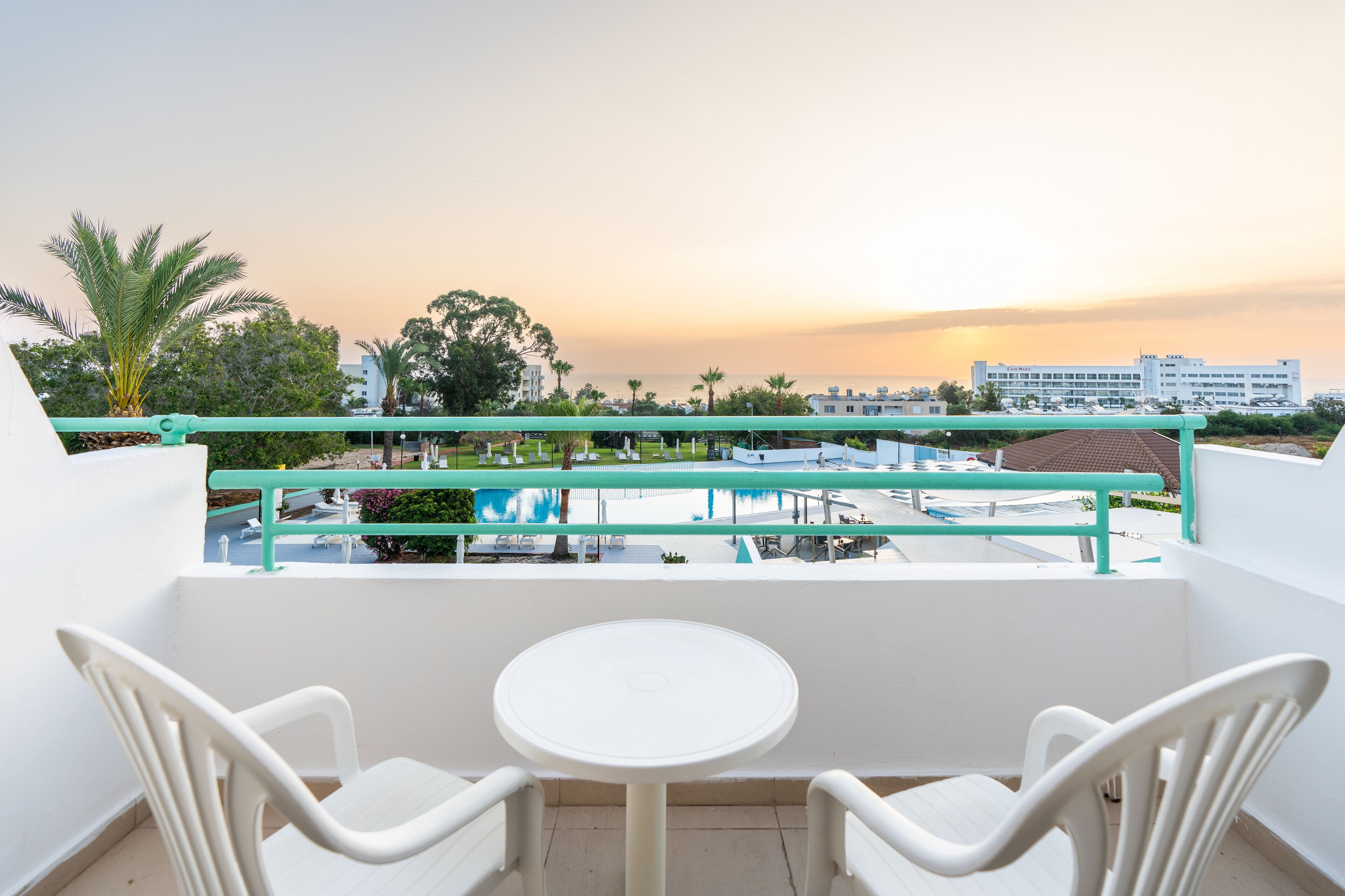 Un balcón con una mesa y sillas blancas, con vistas a una piscina y edificios.