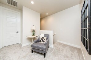 Un pasillo con una puerta blanca, un sillón gris con un cojín, una mesita con una lámpara y una ventana con barrotes.