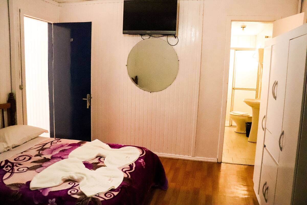 Un dormitorio con una cama, una puerta, un espejo, un televisor y un baño visible a través de una puerta abierta.