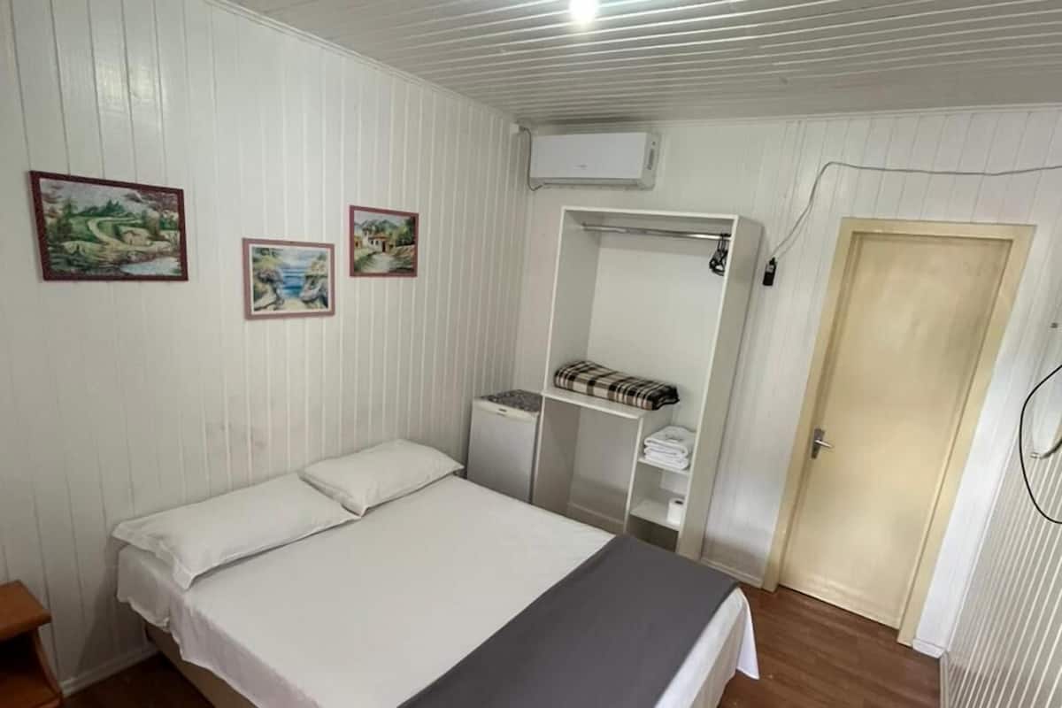 Habitación con una cama individual, suelo de madera, paredes blancas, ropa de cama blanca y gris, una mesita de noche pequeña, un armario y tres cuadros en la pared.