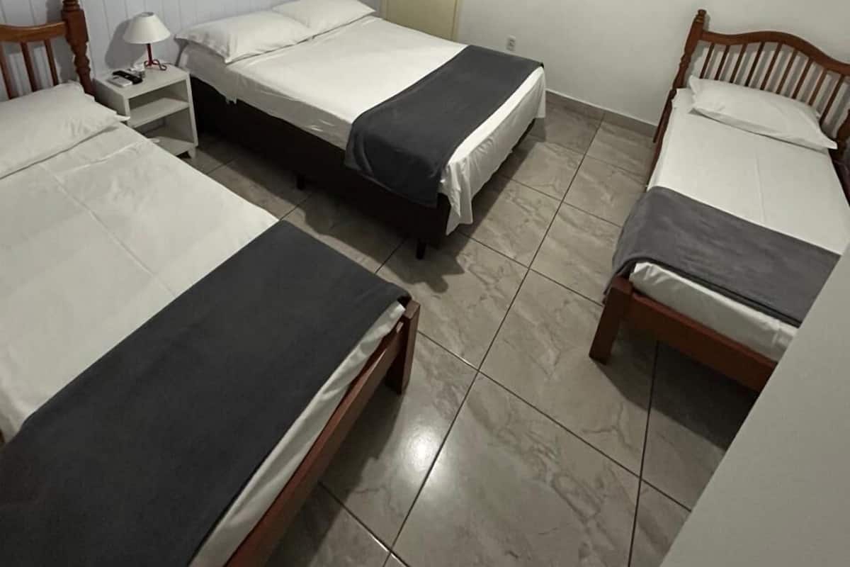 Una habitación con tres camas individuales, una mesita de noche con lámpara, un espejo redondo y dos cuadros de flores colgados en la pared.