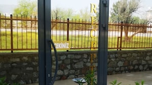 Entrada com porta de vidro, portão de metal, plantas em vasos e parede de pedra ao fundo.