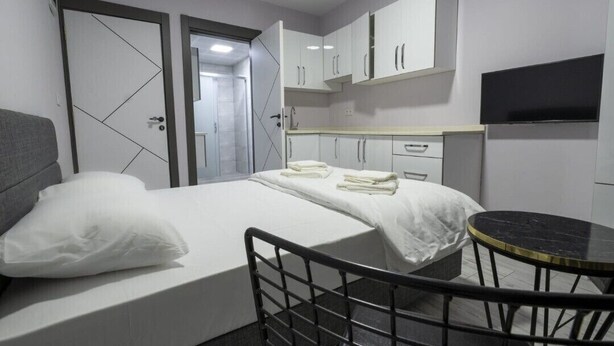 Quarto de hotel moderno com cama, uma pequena mesa redonda, televisão e cozinha compacta.