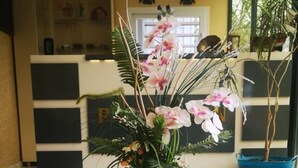 Uma mesa branca com um arranjo floral em vaso branco, sobre um chão de mármore em um quarto com parede amarela e uma janela.