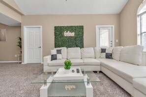 Un salon avec un canapé beige, une table basse en verre ornée d’une plante, et un panneau mural sur lequel on peut lire : « Croyez en vous ».