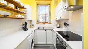 A compact kitchen with white cabinets, a built-in washing machine, a stove, and a window with curtains. - The Tollbooth - The Ideal Base for Exploring (Clackmannanshire)