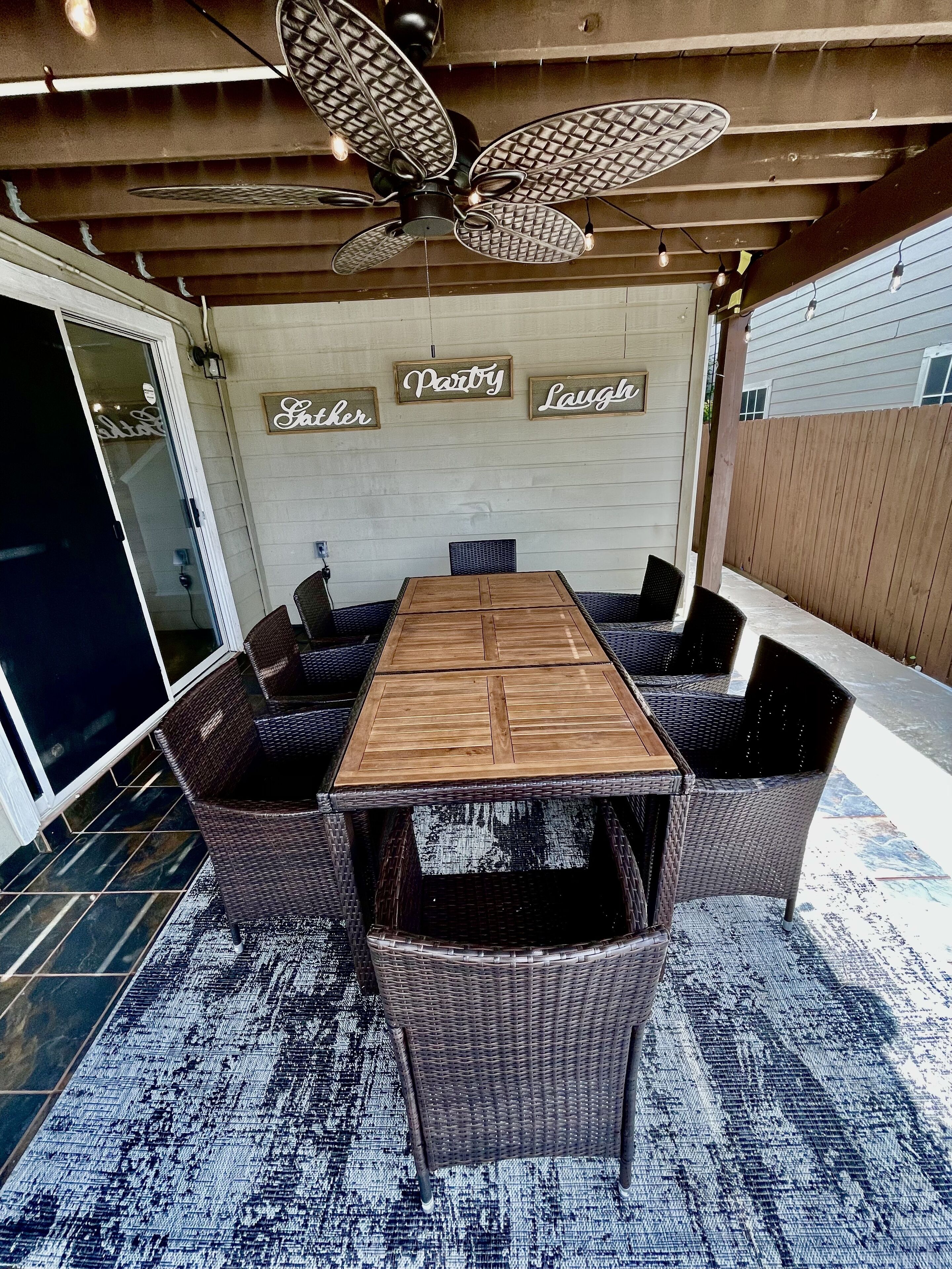 Un patio techado con una mesa de madera y sillas de mimbre, un ventilador de techo y carteles decorativos en la pared.