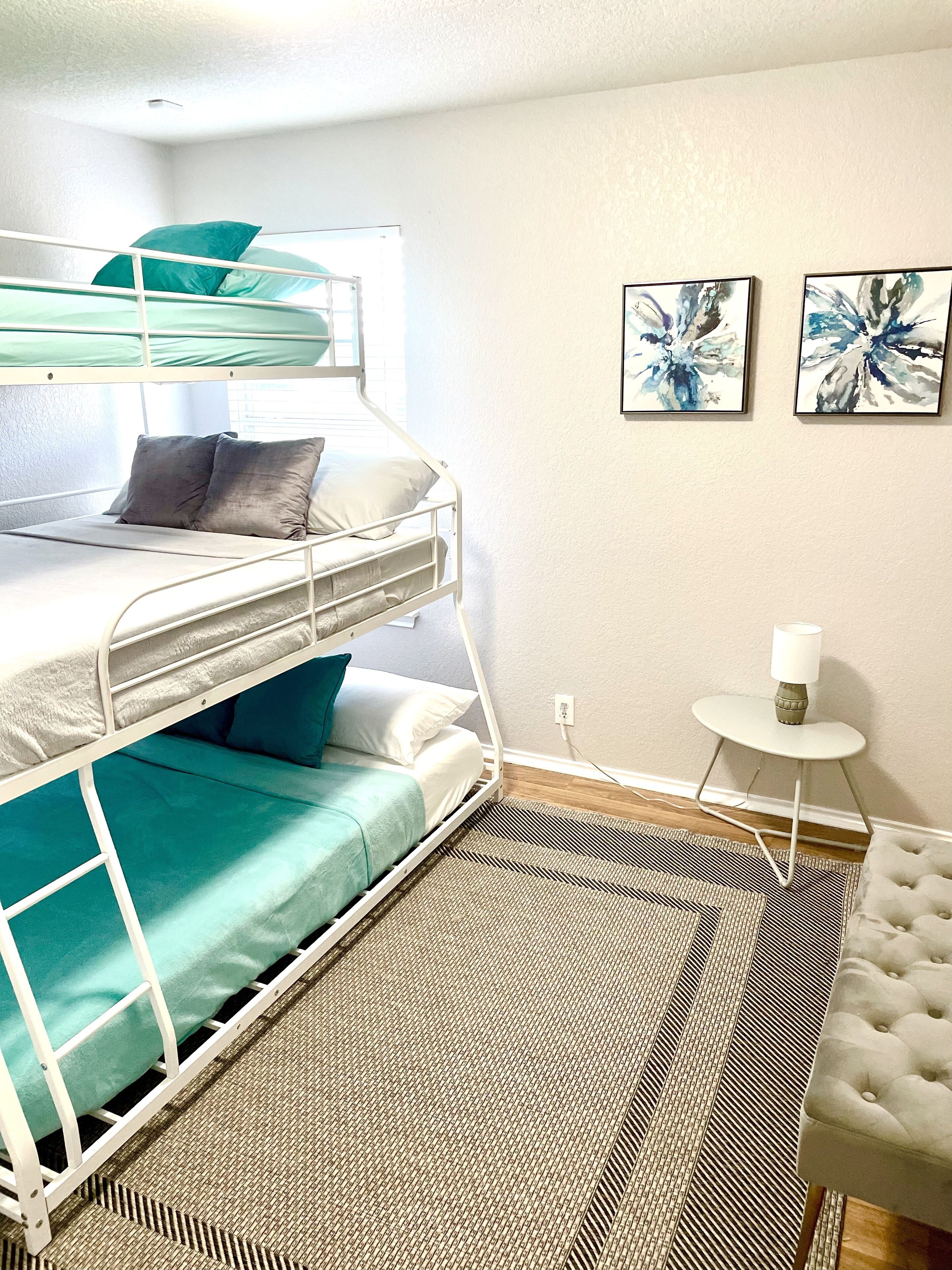 A bunk bed with teal and gray bedding, a small white table with a candle, and two abstract paintings on the wall.