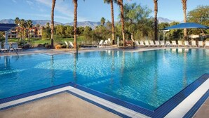 A swimming pool with lounge chairs and palm trees, surrounded by a landscaped area with mountains in the background. - Coachella 2023 (Palm Desert)