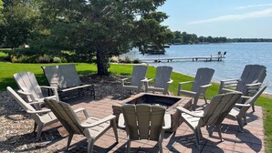 Un espacio al aire libre con sillas Adirondack y una hoguera, con vista a un lago.