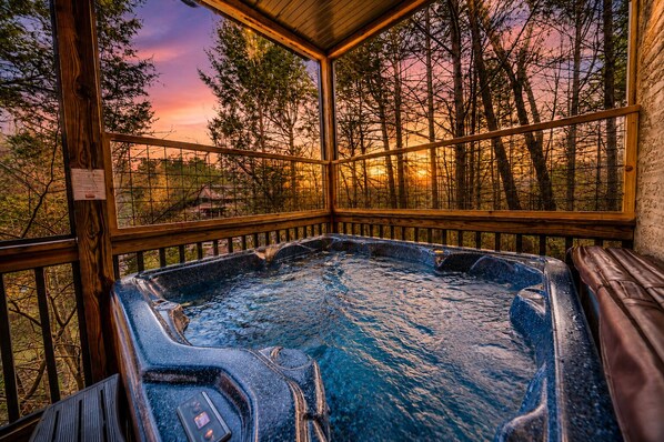 A hot tub with a wooden deck and glass walls, surrounded by trees and a sunset view.