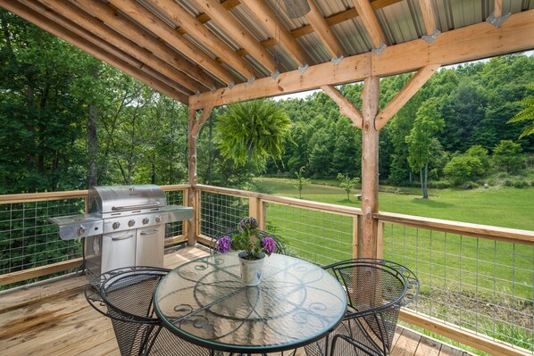 An outdoor patio with a grill, a round table with chairs, and a view of a green landscape. - Creekside Cabin, located just minutes from Blowing Rock! (Lenoir)