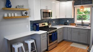 A kitchen with white cabinets, a stainless steel stove, and a microwave. There are two white stools and a window with a view of greenery. - Pristine condition! Three bedroom sleeping 8 with contemporary beach decor! (South Dennis)