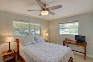 A bedroom with a bed, two bedside tables with lamps, a ceiling fan, a TV on a wooden desk, and windows with a view of greenery.