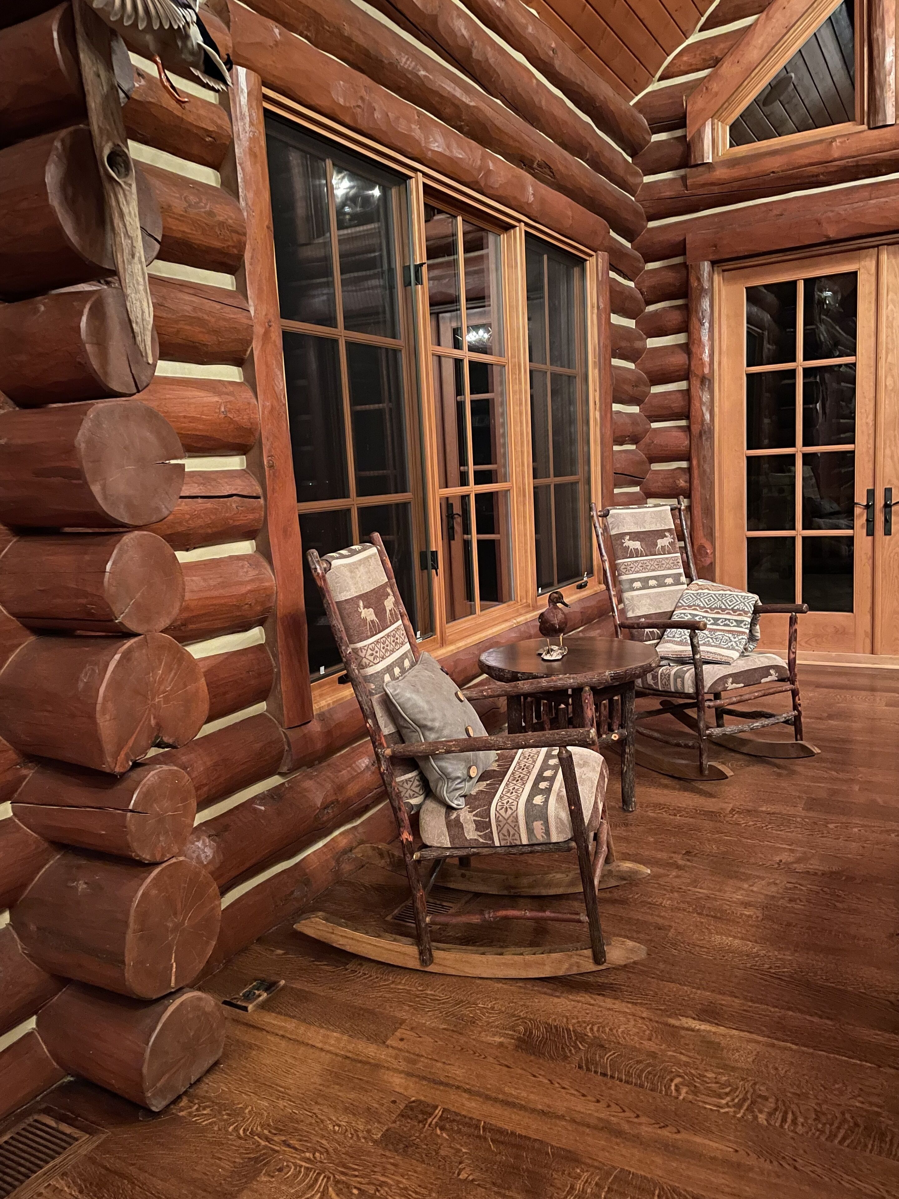 A log cabin interior with a wooden floor, a round table, two upholstered chairs, and a large window with a view of the outside.