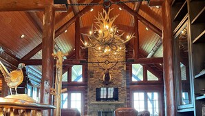 Un intérieur de chalet spacieux, agrémenté d’une cheminée en pierre, de fauteuils en cuir et d’un lustre rustique.