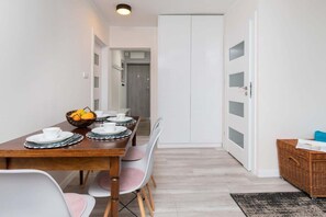 Et spiseområde med et træbord dækket til fire personer, hvide stole, en kurv med frugt og en hvid garderobe med glasdøre med sandblæsning.
