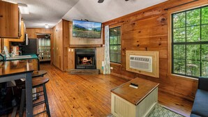 A cozy cabin interior with wooden walls, a fireplace, a ceiling fan, and a large window offering a view of the outdoors. - Romantic Cabin w/ Sunset Views • Hot Tub • Near Gatlinburg (Sevierville)