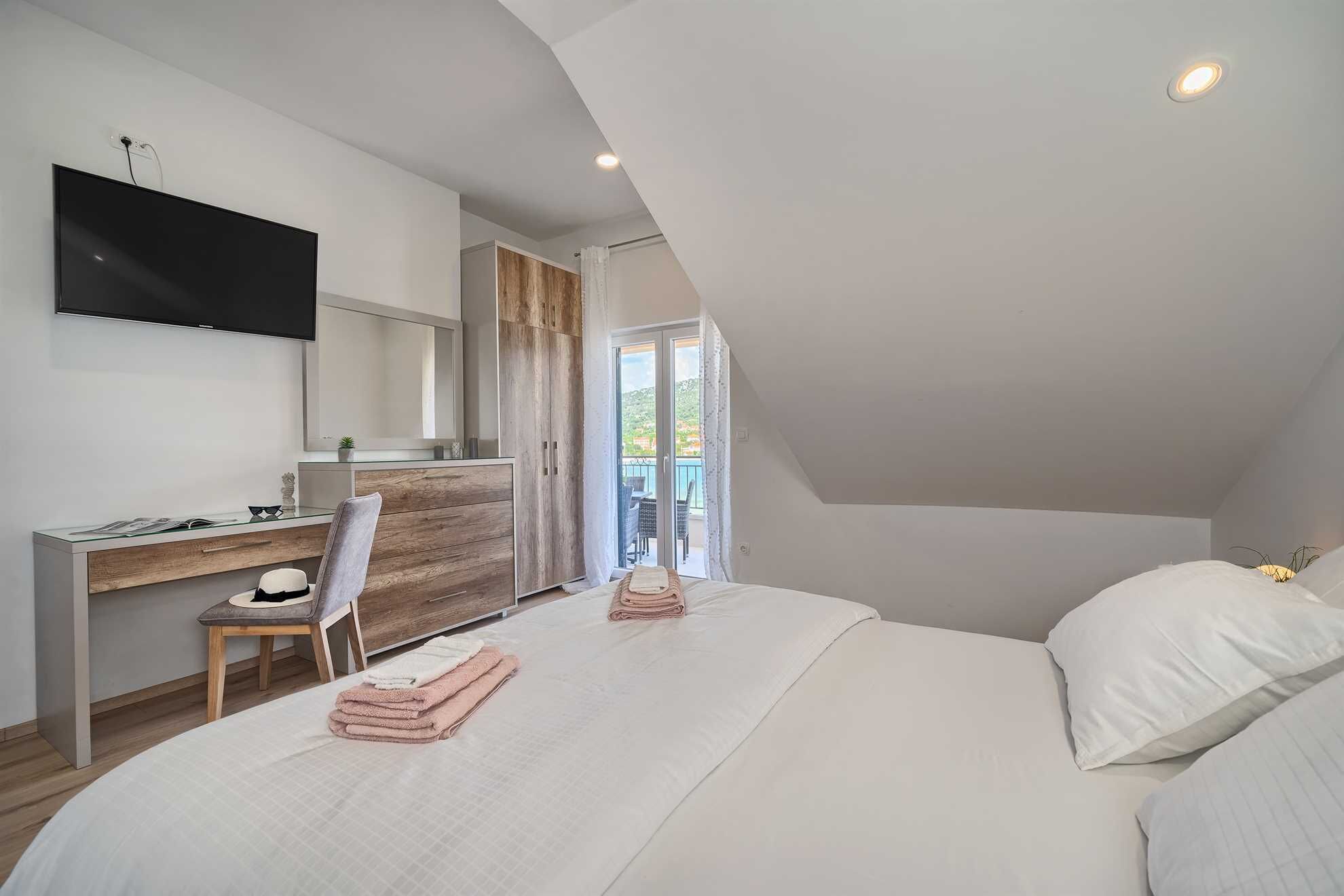 Un dormitorio moderno con una cama grande, un escritorio y un televisor. Hay un balcón con vistas al exterior.