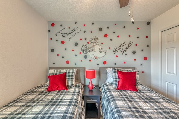 A bedroom with two beds, each with plaid bedding and red pillows, a nightstand with a red lamp, and a wall decorated with red and gray polka dots and a Mickey Mouse design.