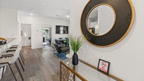 A modern living room with a large round mirror, a dining area with bar stools, and a staircase with a 'No Smoking' sign. - A · New 4Br King Beds Steps to Beach &2nd Pier #A (Myrtle Beach)