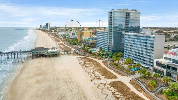 A coastal cityscape with a beach, a pier, and modern buildings including a hotel with a glass facade.