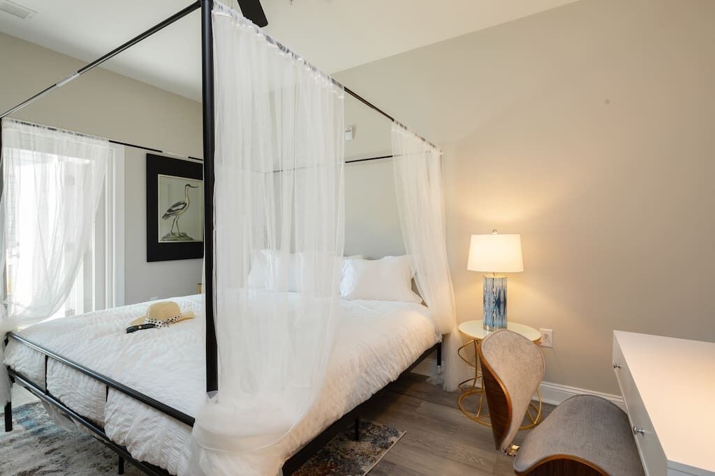 Una cama con dosel y cuatro columnas, con una colcha blanca, una mesita de noche con lámpara y una silla con cojín.