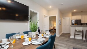 Un comedor con una mesa larga dispuesta para el desayuno, un televisor montado en la pared y una cocina al fondo.