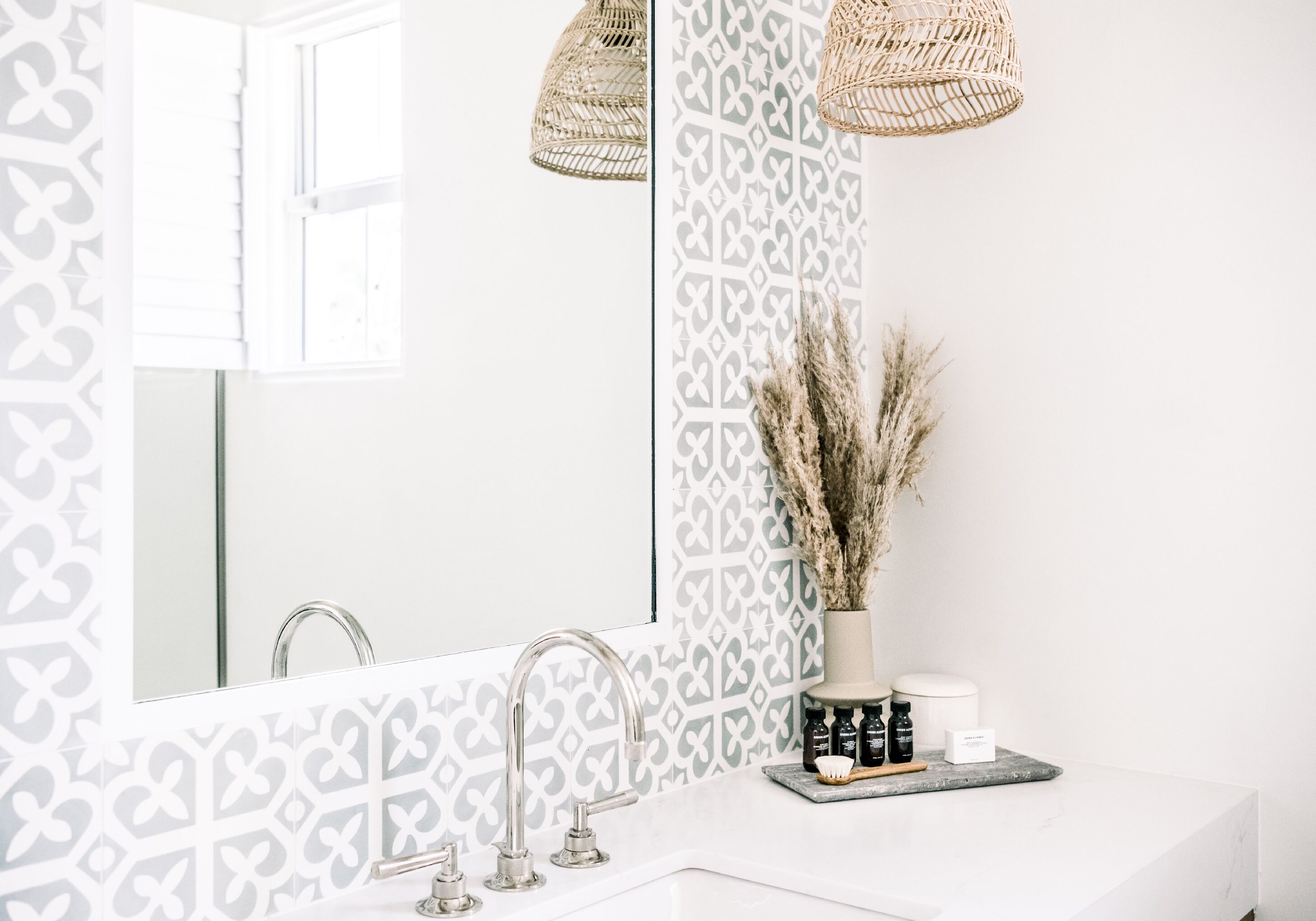 Un baño con un lavabo blanco, un espejo grande y una pared con diseño. Se ven dos lámparas de tela de mimbre sobre el espejo.