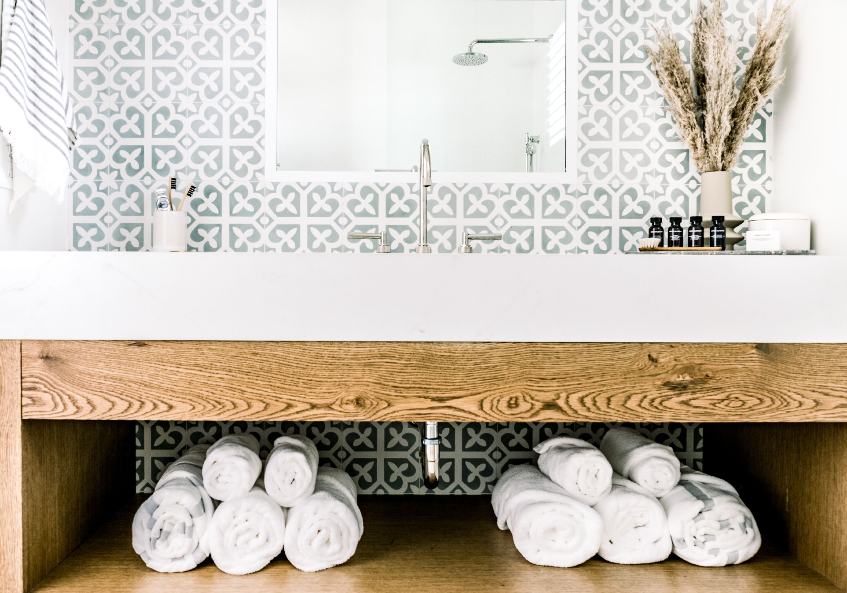 A bathroom with a patterned backsplash, a mirror, a sink with a faucet, and a wooden shelf below holding rolled towels.