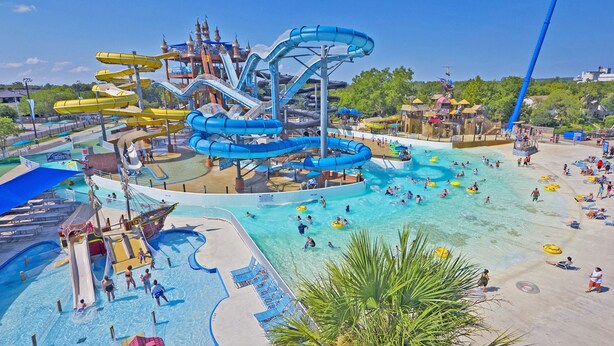 A water park with multiple slides and pools, including a large central water slide, smaller slides, and a sandy beach area with lounge chairs.