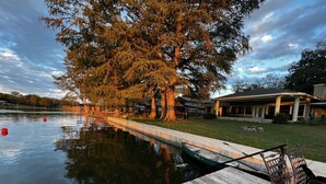 A wooden dock extending into a calm lake, with a large tree and a building in the background.