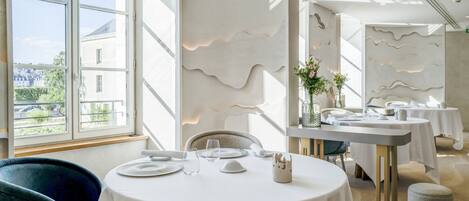 Un comedor con mesas redondas, manteles blancos y vistas a un edificio a través de amplios ventanales.