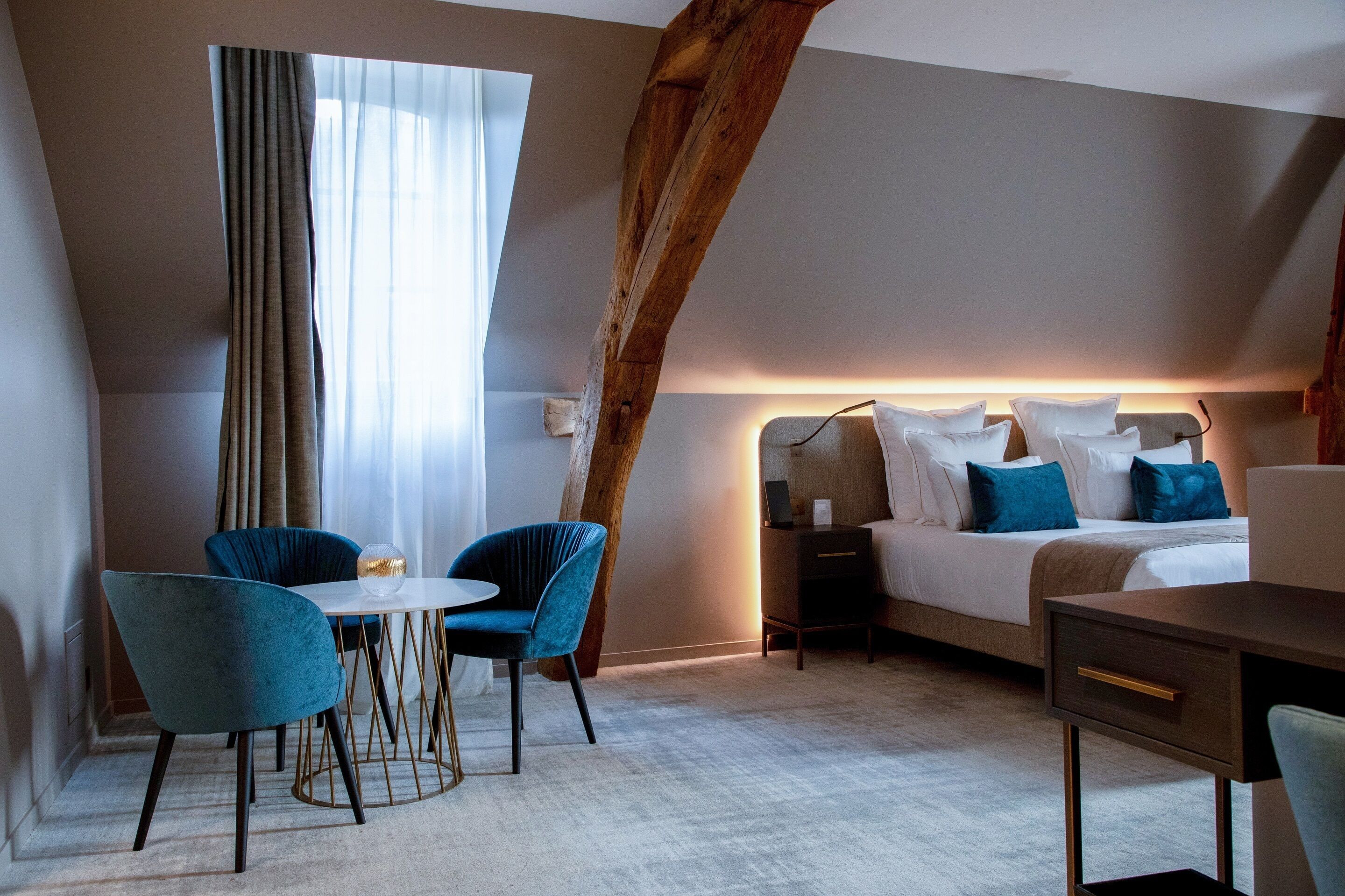Une chambre d’hôtel moderne avec un plafond de style industriel, un lit avec du linge de lit blanc et des oreillers bleus, un bureau avec une chaise, et un coin repas avec des chaises bleues et une table ronde.