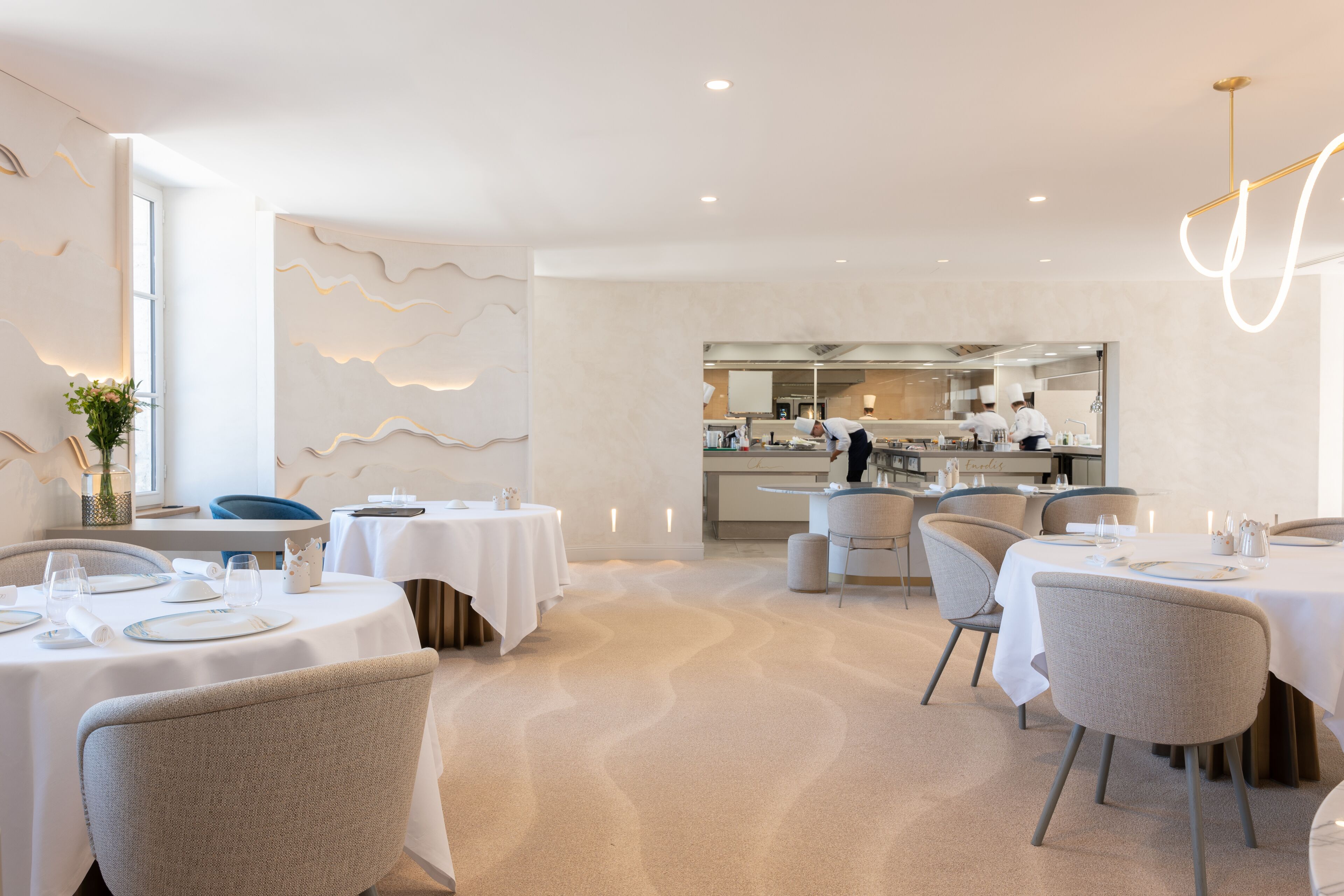 A modern dining area with round tables set for four, a kitchen in the background, and a wavy wall design.