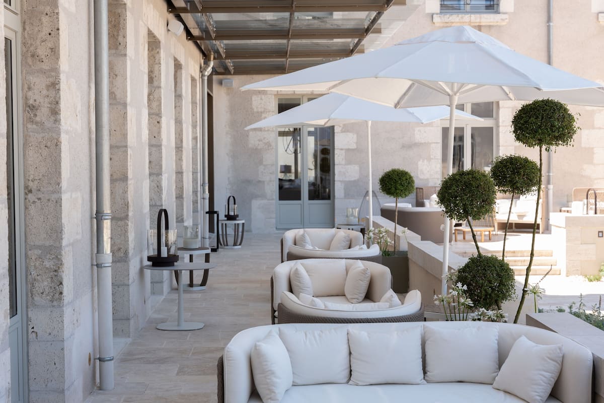A modern outdoor seating area with white sofas and umbrellas, a stone wall, and a glass door leading to an interior space.
