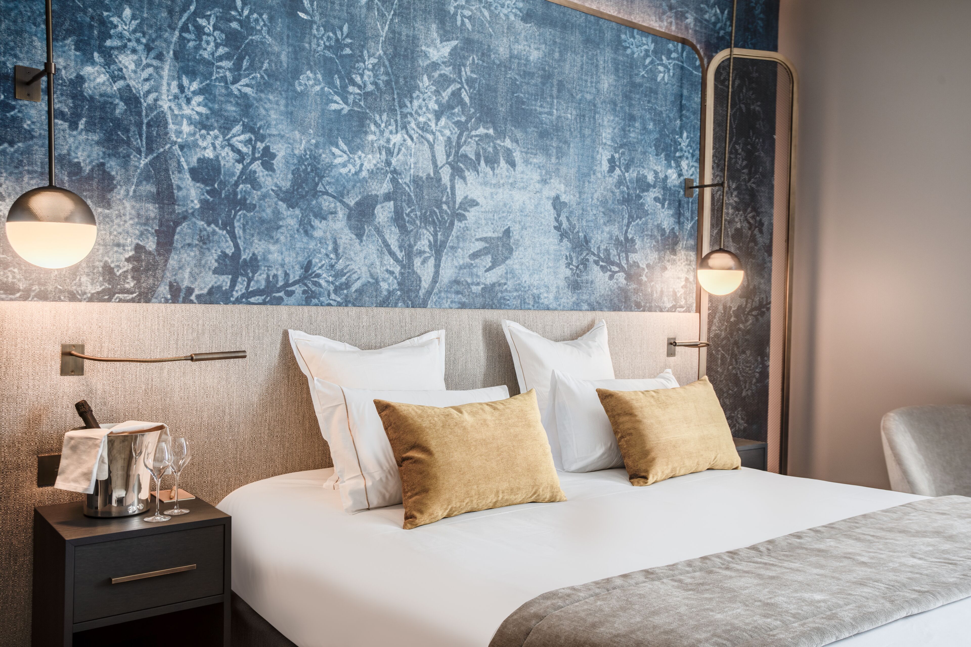 Una cama bien hecha con almohadas blancas y color beige, una mesita de noche con una lámpara y una pared con estampado floral.