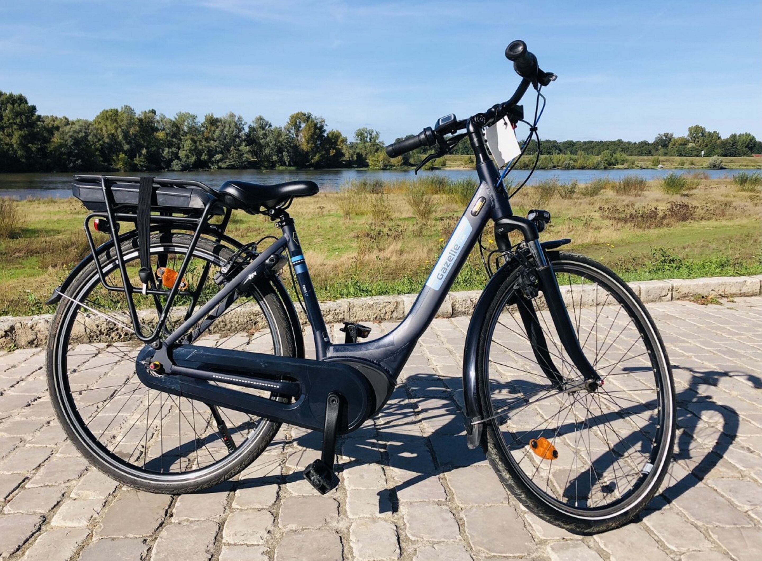 En stillastående elcykel med en korg fram och ett bagageplan bak, parkerad på en kullerstensbelagd stig vid en sjö.