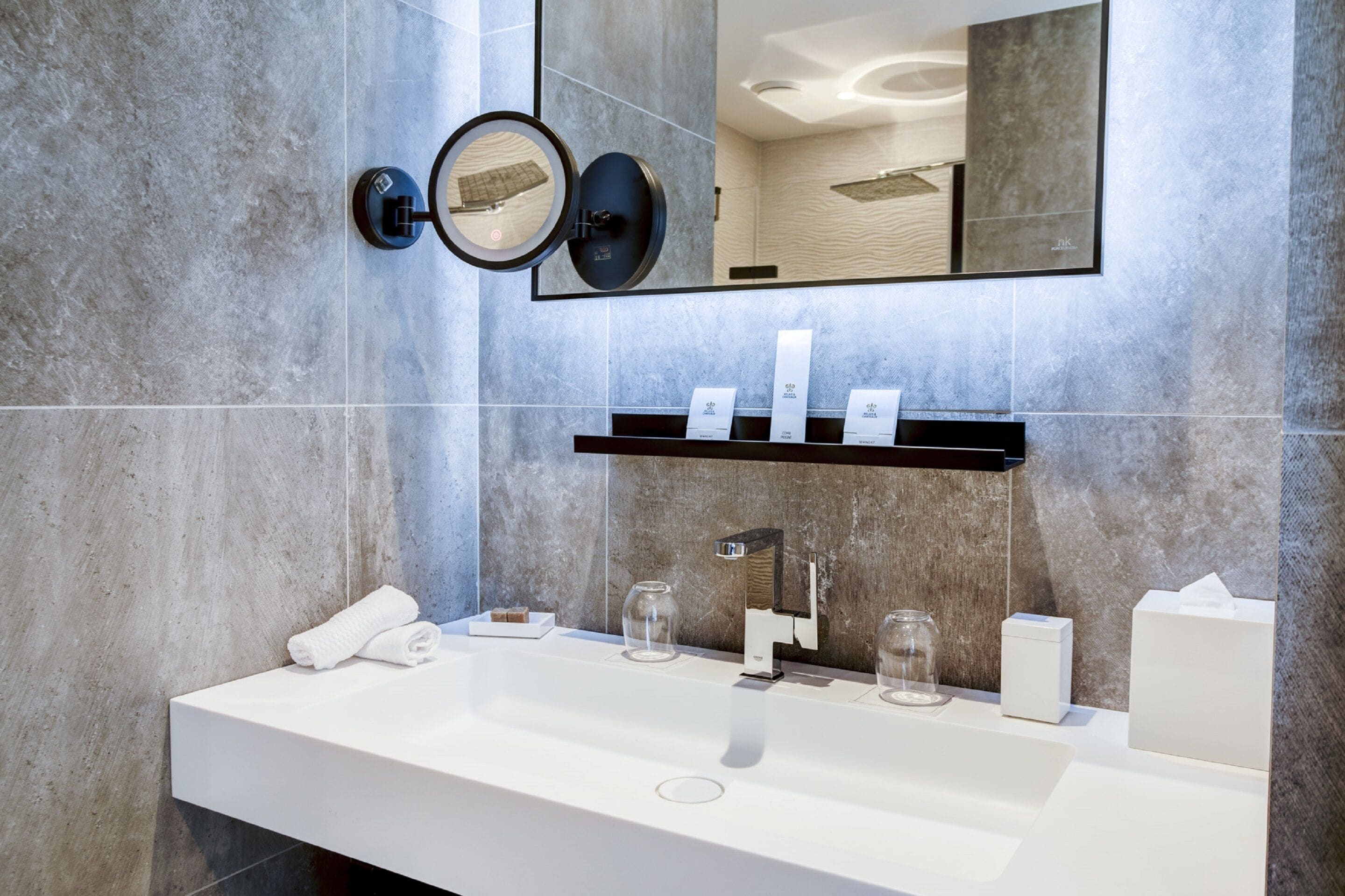 A modern bathroom with a large mirror, a white sink, and a shelf with toiletries.