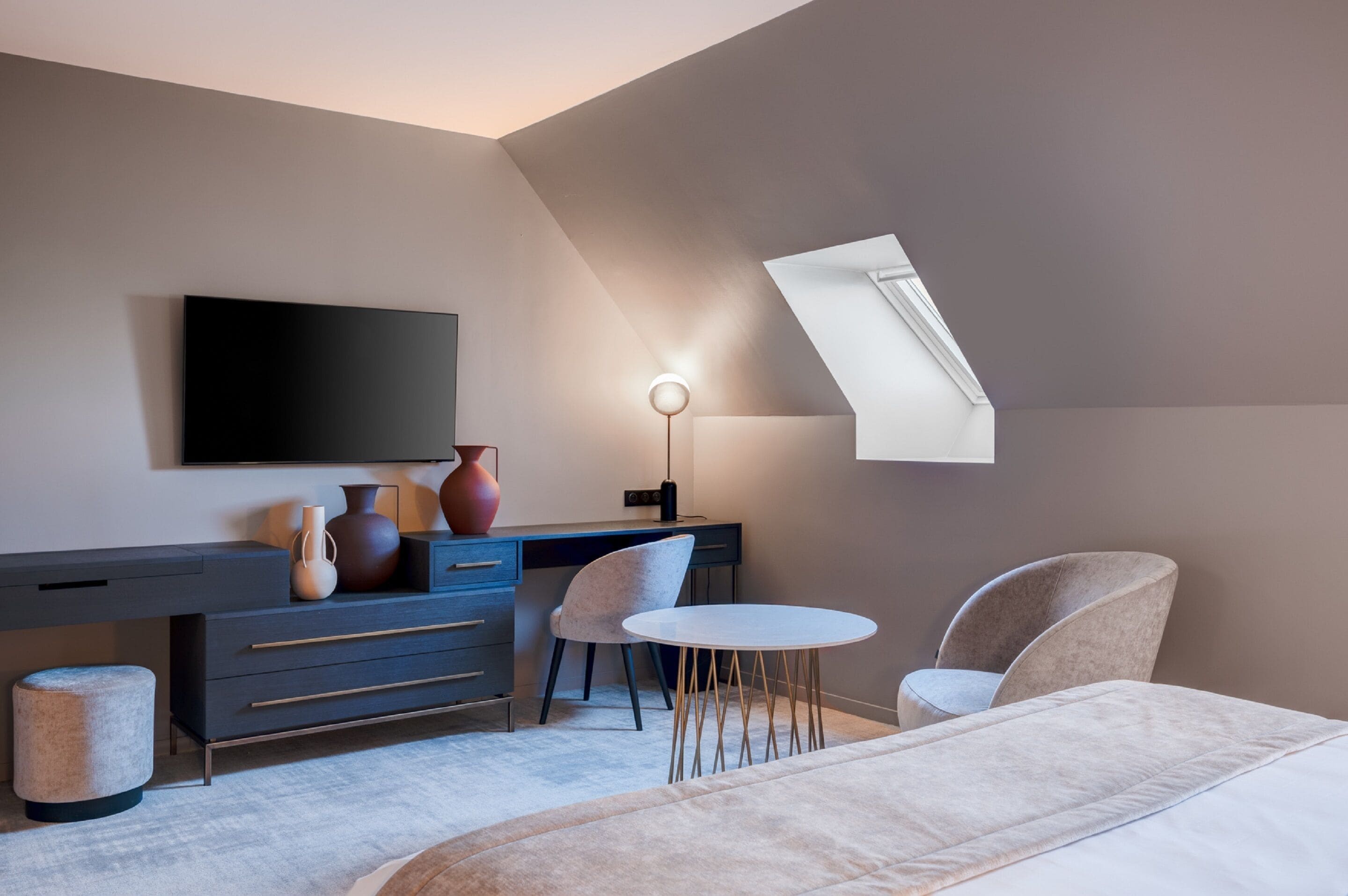 Une chambre moderne équipée d’un bureau, de deux chaises, d’un lit, d’une télévision et d’une fenêtre de toit.