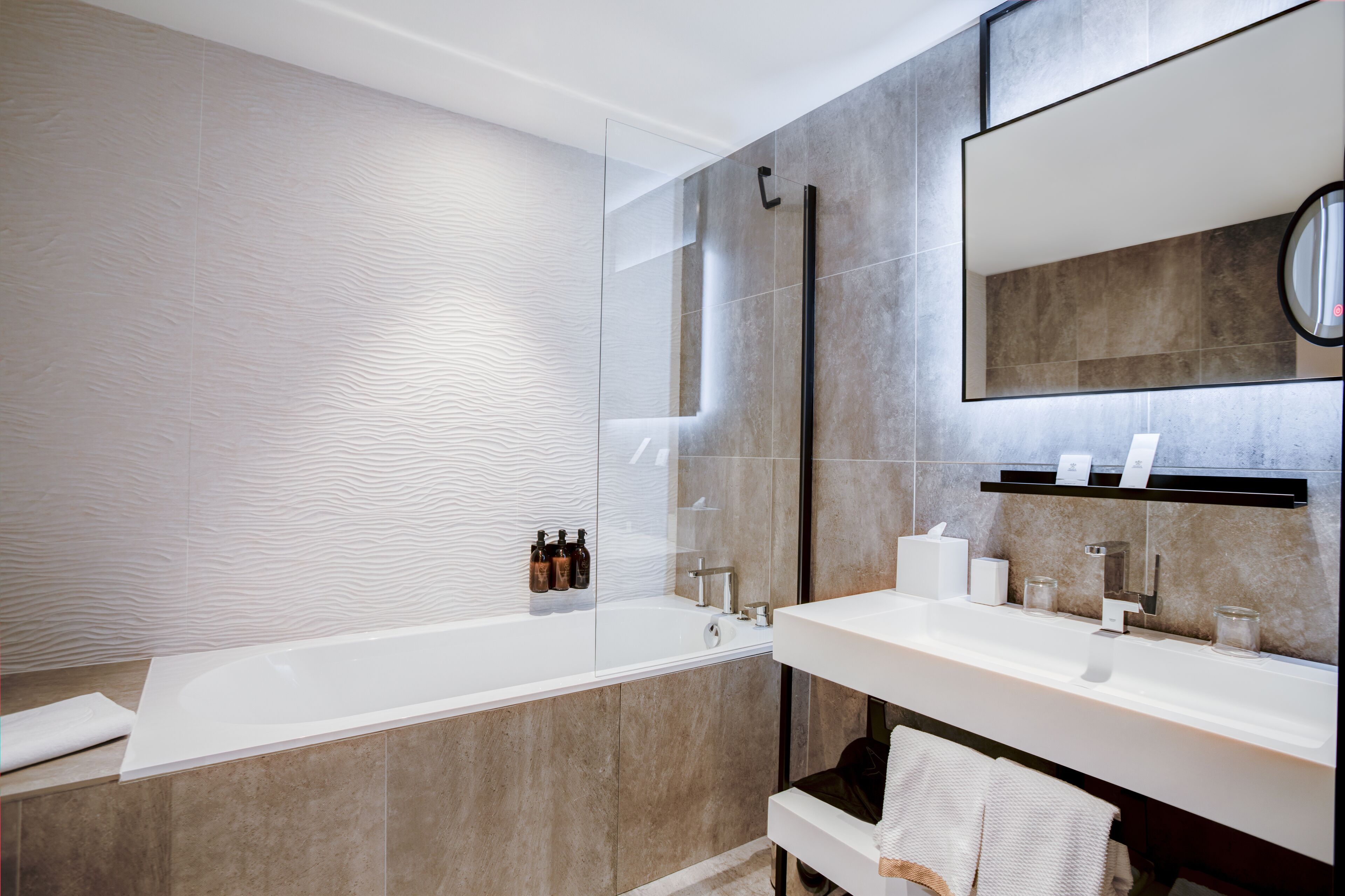 Une salle de bain moderne avec une baignoire entourée de parois en verre, un grand miroir et un meuble-lavabo doté d’un porte-serviettes.