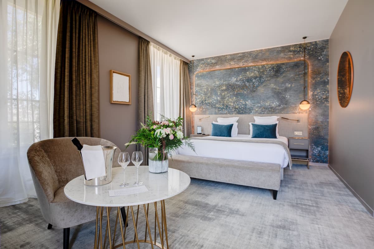A hotel room with a large bed, a bedside table with a vase of flowers, a chair, and a round table with wine glasses.