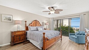 Ein Schlafzimmer mit einem großen Bett, einem Holzkopfteil, zwei Nachttischen, einem Beistelltisch, einem blauen Sessel und einem Balkon mit Blick.