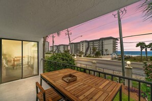 Un balcon avec une table et des chaises en bois, donnant sur une rue et des immeubles, et offrant une vue sur l’océan au coucher du soleil.