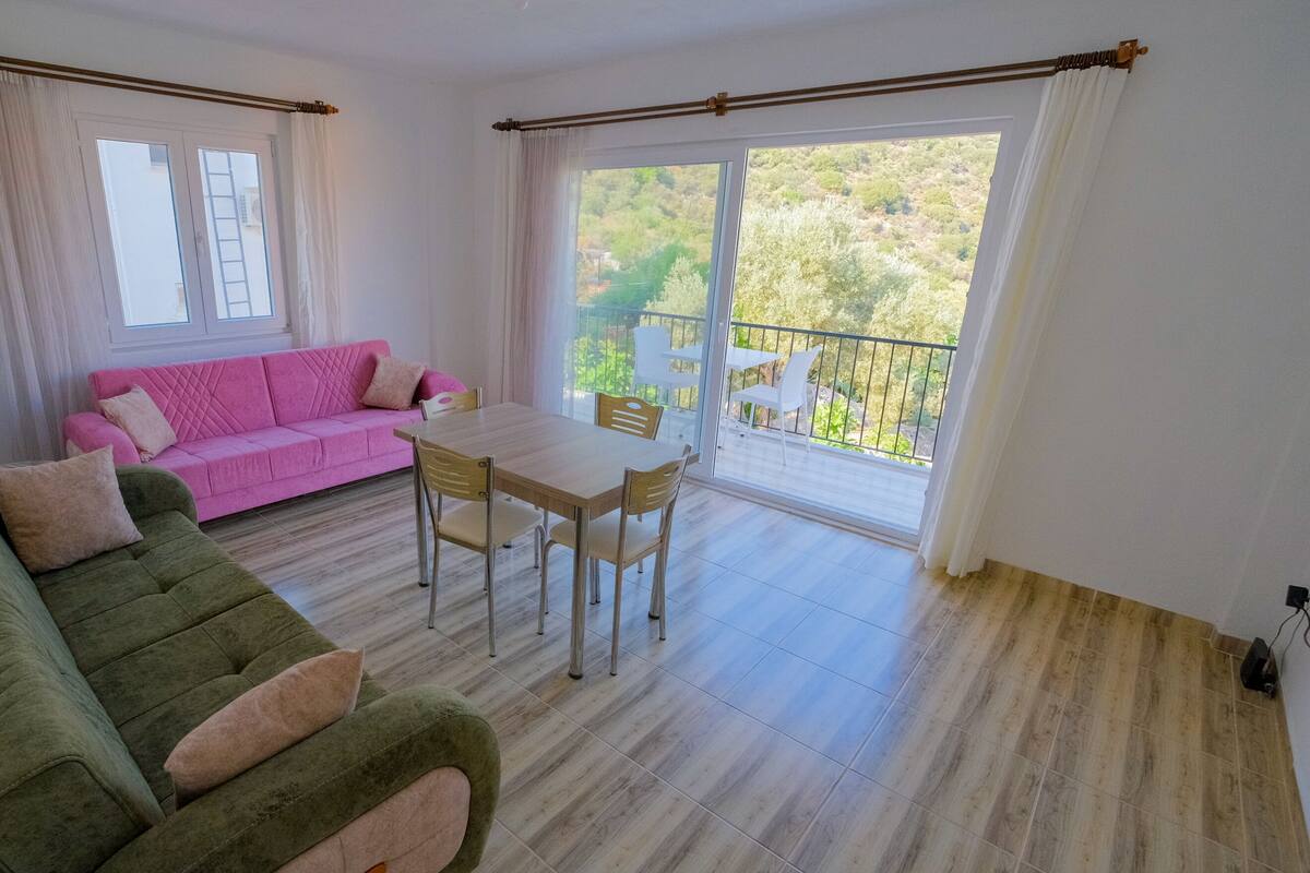 A living room with a green sofa, a pink sofa, a wooden dining table, and chairs. There is a balcony with a view of greenery.