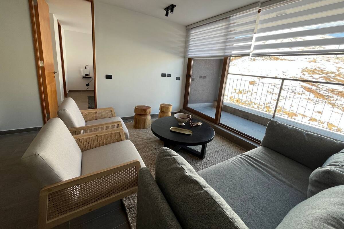 A modern living room with a sofa, two armchairs, a coffee table, and a large window with blinds.