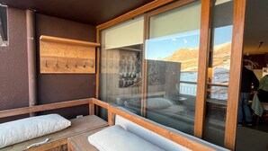 A wooden deck with a bench and cushions, a wooden coat rack with hooks, and a large window with a view of a mountainous landscape.